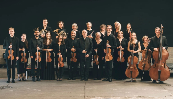 The players in black concert dress stand in two rows whilst holding their instruments